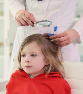 Pediatrician looks for lice on head of little girl sitting in chair. Doctor checks hair of little patient using magnifier glass in clinic