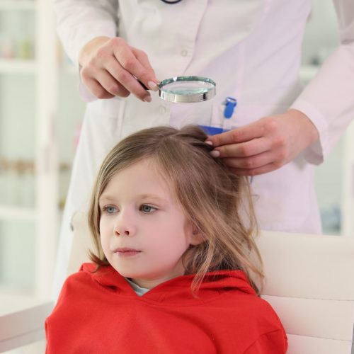 Pediatrician looks for lice on head of little girl sitting in chair. Doctor checks hair of little patient using magnifier glass in clinic